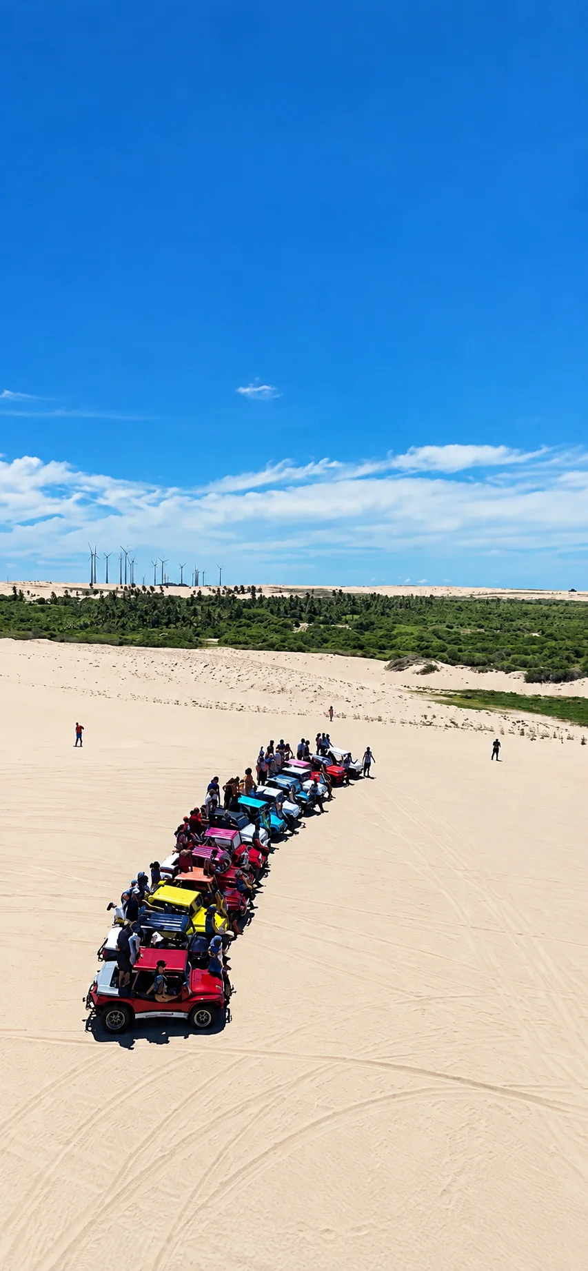 Fila de buggys sobre as dunas, com aerogeradores ao fundo